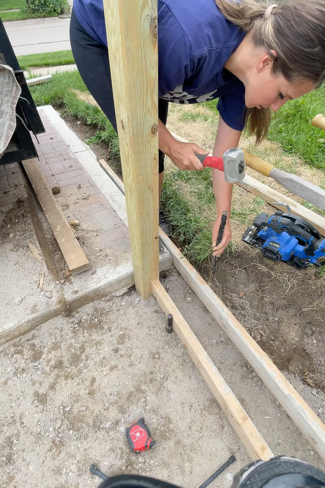 Woman hammering rebar into the ground to secure wood framing for a DIY concrete curb to border a small paver patio.